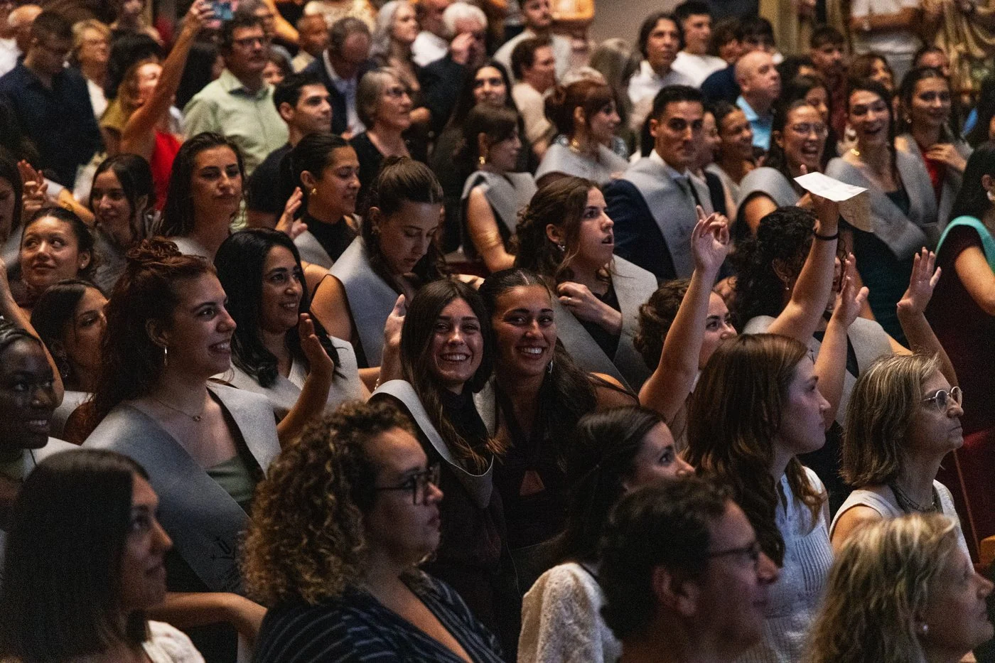 Dos graduadas sonríen a la cámara entre el público durante el acto de graduación