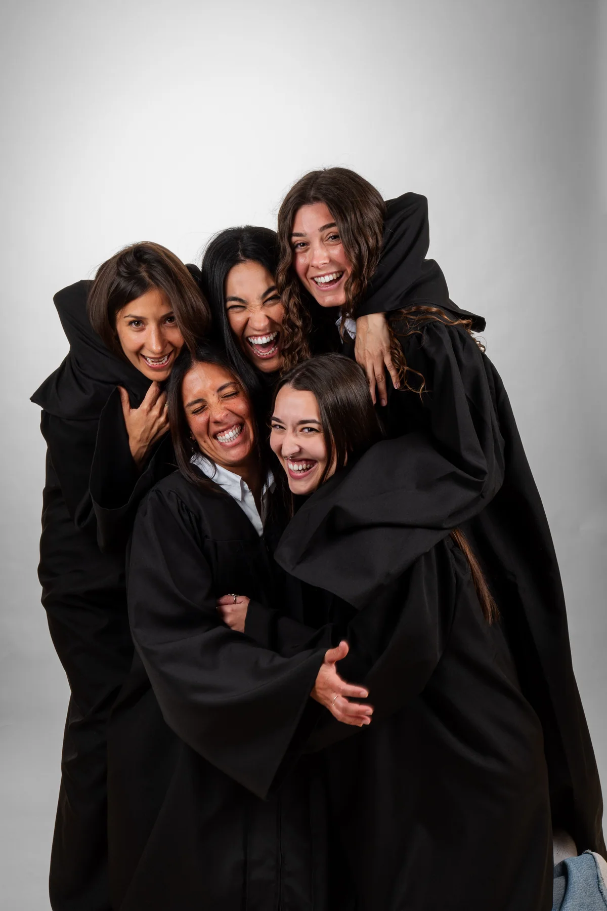 Cinco graduadas con toga se abrazan riendo en el estudio