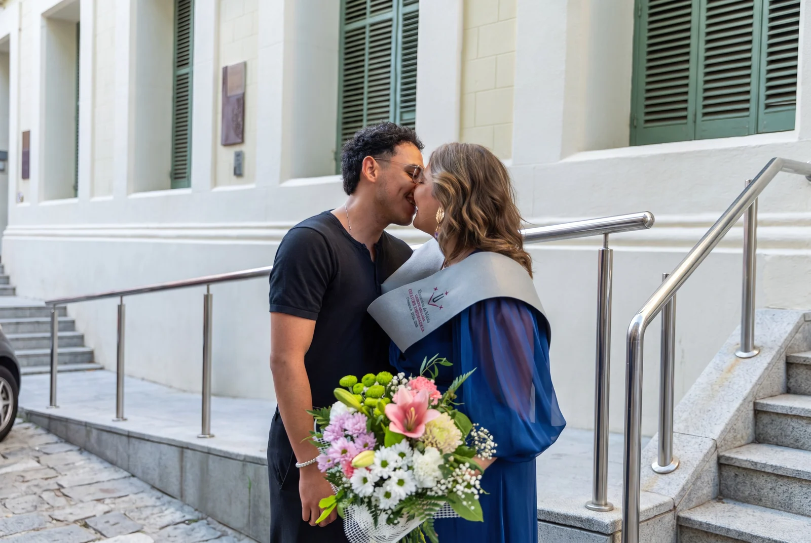 Una graduada con banda y ramo de flores se besa con su pareja a la salida del acto