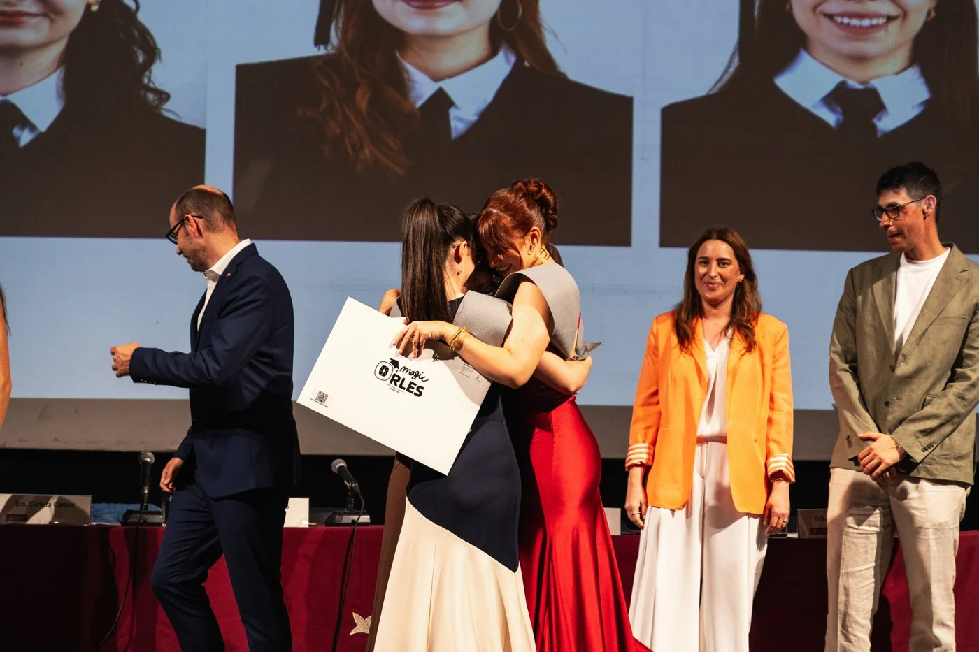Una graduada con vestido rojo abraza a una compañera sobre el escenario con el sobre de MagicOrles en la mano
